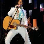 Elvis tribute artist JD King on stage in white jumpsuit holding guitar